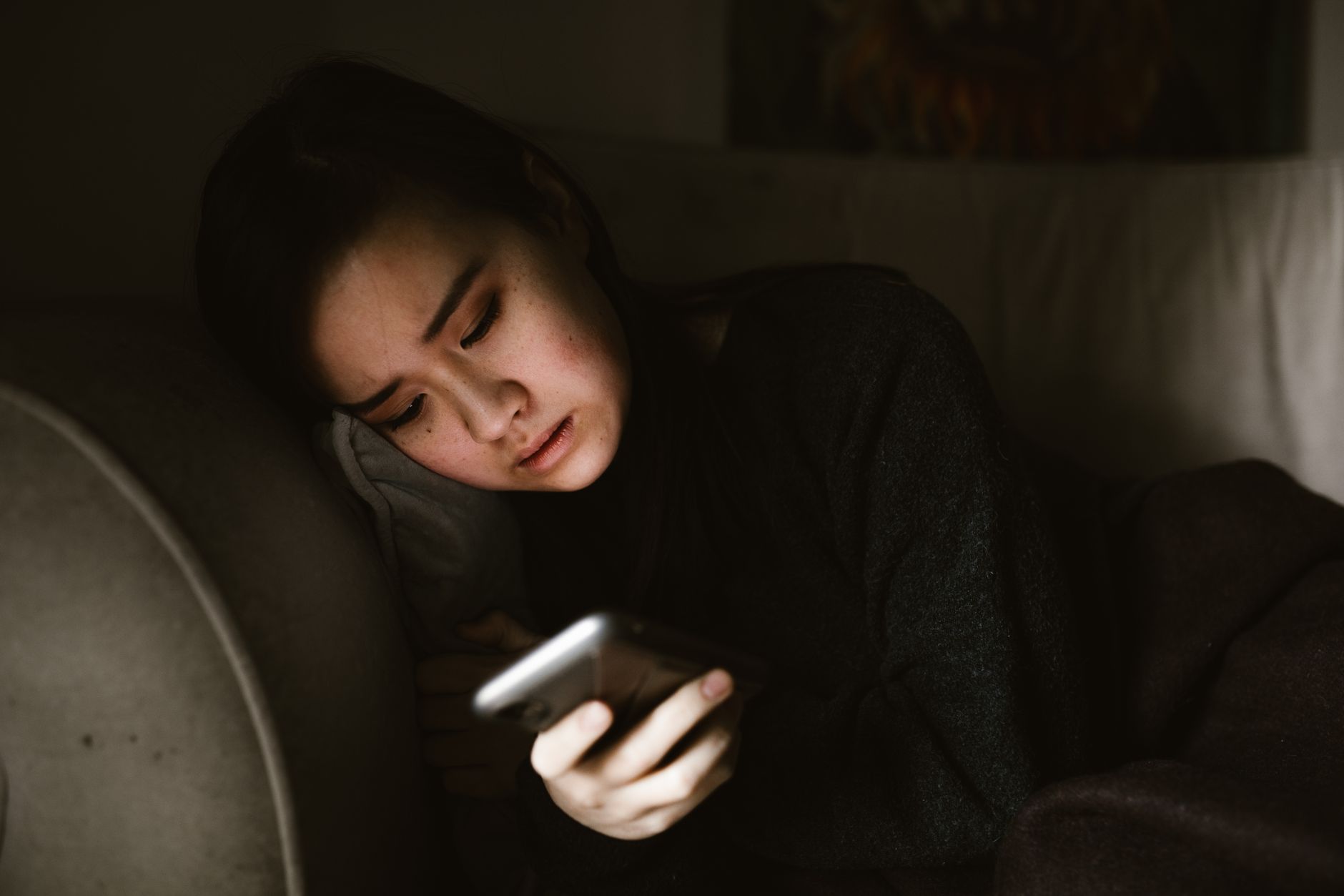 Woman on a couch in a dimly lit room holding a smartphone, looking worried at a celebrity profile on the screen