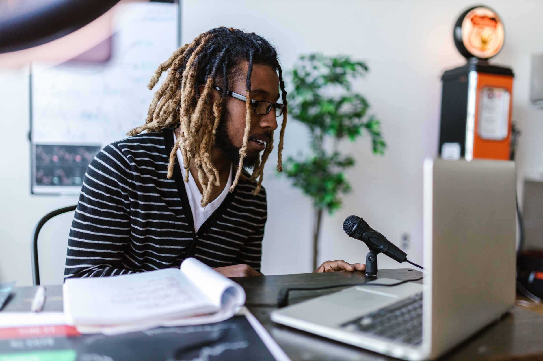 Man at a desk wearing headphones and speaking into a studio microphone while a laptop shows audio editing software